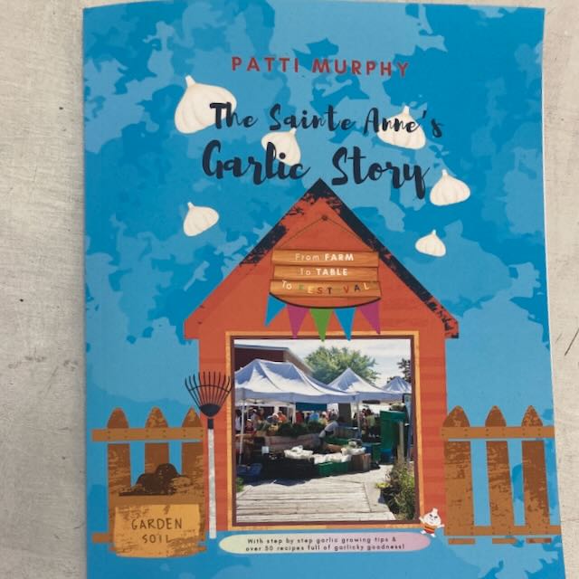 La couverture bleue d&#39;un livre de TS intitulé « Livre - The Sainte Anne&#39;s Garlic Story » de Patti Murphy comporte une illustration d&#39;un hangar en bois avec une fenêtre représentant une scène de marché. Sous le titre, on peut lire, en texte anglais, « From Farm to Table Festival ». La couverture comprend également des illustrations d&#39;ail et des graphiques de terre de jardin.