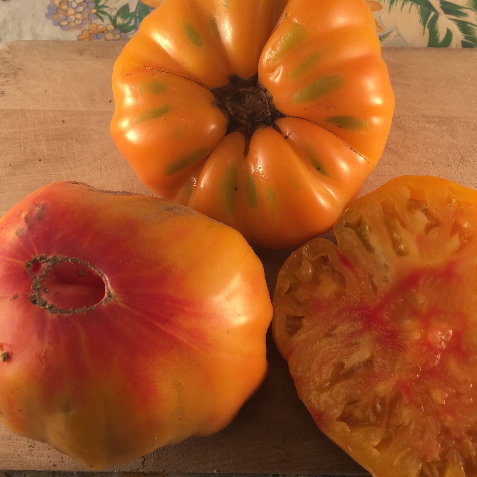 Trois tomates Orange Striped de Tourne-Sol, de couleur orange et rouge, reposent sur une surface en bois. L'une d'entre elles a été tranchée pour montrer son intérieur juteux. Un tissu à motifs floraux est visible à l'arrière-plan.