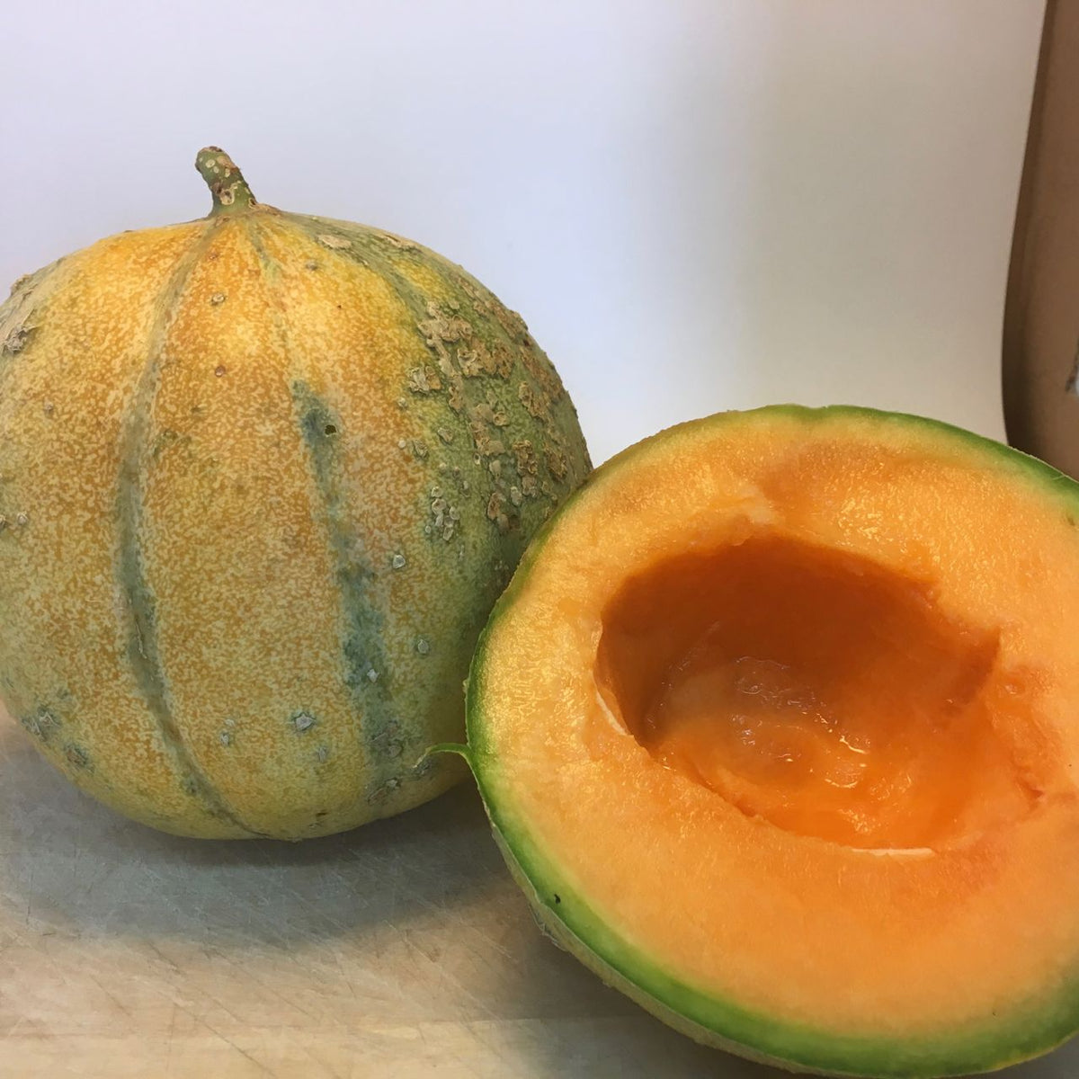 Un Melon Petit Gris de Renne entier de Tourne-Sol, à la peau texturée verte et jaune, repose à côté d&#39;une moitié présentant une chair orange vibrante et un centre creux, révélant son goût riche et sucré.