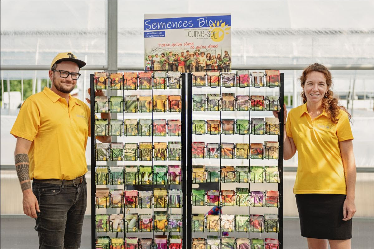 Deux personnes en chemise jaune se tiennent à côté d'une exposition colorée de Don de Semences. Le panneau indique « Tourne-Sol » et le décor intérieur a une atmosphère de serre, soulignant l'aspect organique.