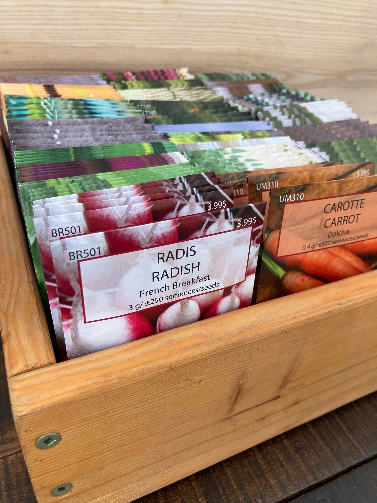 Les sachets de la « Collection Fermiers - C - 15 variétés x 10 sachets » de Tourne-Sol, contenant des graines de légumes populaires comme le radis et la carotte, sont soigneusement disposés dans une boîte en bois. Les étiquettes indiquent la quantité et le prix des graines sur des planches de bois rustiques, idéales pour les jardiniers à la recherche de semis directs.